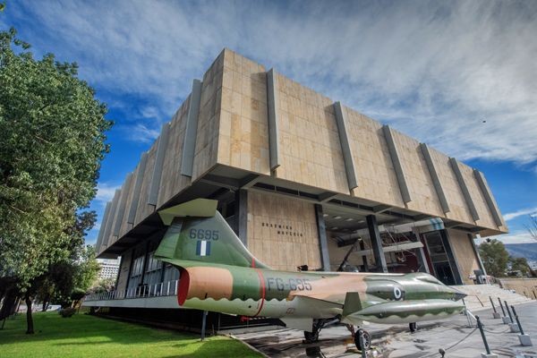 Exterior view of a large, modern museum building with a vintage military aircraft displayed in front.