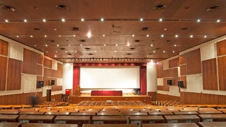 Empty auditorium with rows of wooden seating facing a large screen and stage with red curtains.